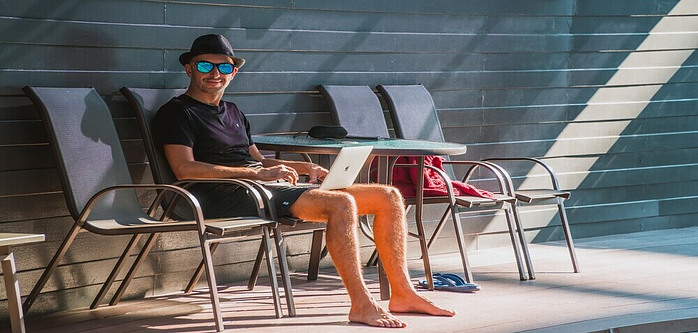 A Gentleman sitting in the Sun, wearing a hat and Sunglasses. On his Lap he has an open Laptop as he types. The image is to convey writing a blog post as per the article Why Consider Starting a Blog by Future You Success