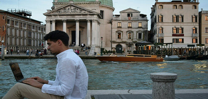 Man on left hand side in the foreground, sitting on a pavement with the backdrop of a Venice canal.