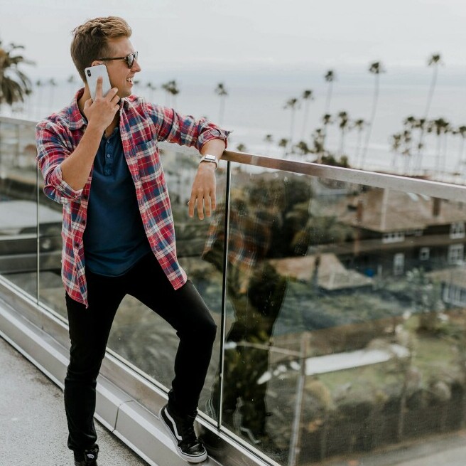 A Young Man in casual clothing is on a balcony with a glass screen. He has his left leg up on the foot rail and he his turned three quarters to us. He is on his mobile phone whilst surveying the view from the balcony, which is a view of a beachfront