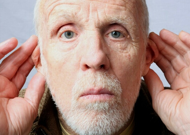 An older male staring straight at the viewer with both his hands cupping his ears as he appears to be struggling to hear what you are saying to him.