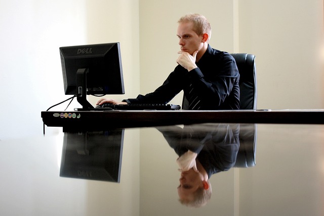 A man sitting at a polished glass office desk looking at his laptop. The polished glass means we can see his reflection perfectly mirroring him in the desk surface