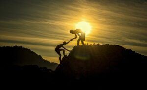 sunset- with a silouetted Man on top of a Mountain helping another man climb up