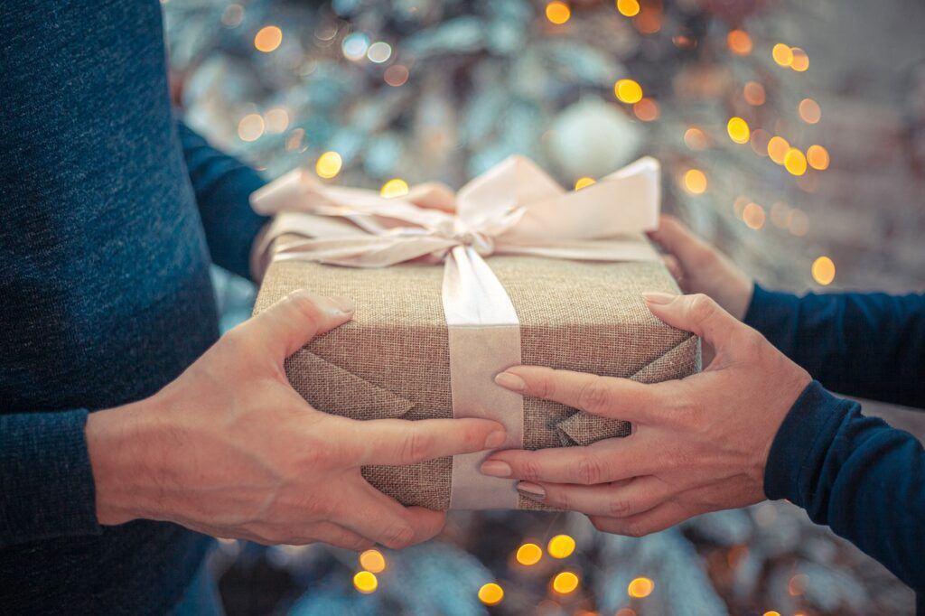 An image of hands belonging to two people. One on the left one on the right. Both have hold of a wrapped gift that is centre image