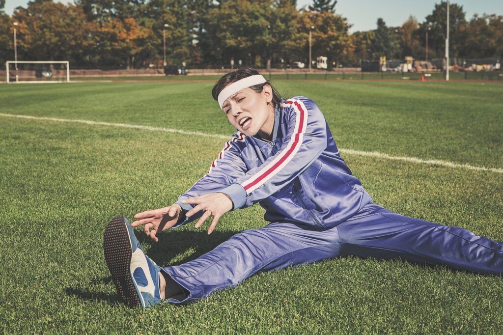 A woman in a lilac tracksuit sitting on the floor with her right leg outstretched. The woman is attempting to  stretch to reach her right foot and grimacing as she does so