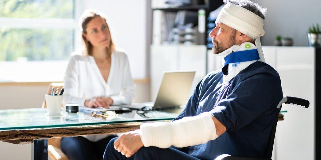 A Man Wearing a Head Bandage and having his left arm in plaster, sitting in front of a Doctor